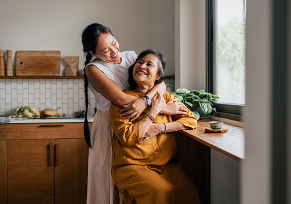 A woman is hugging her mom, who is sitting in the kitchen.